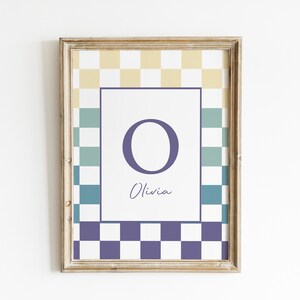 May include: Framed print featuring the letter "O" in a dark purple hue, with the name "Olivia" in cursive below. The print has a checkered pattern in yellow, teal, and purple, and is encased in a gold-toned wooden frame.