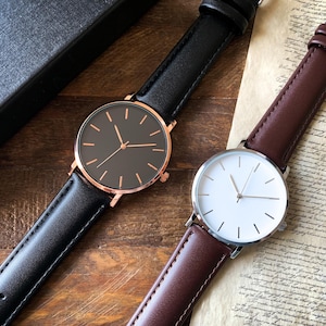 May include: Two minimalist wristwatches. One has a black face with a rose gold case and a black leather band. The other has a white face with a silver case and a brown leather band. Both have simple stick hour markers.