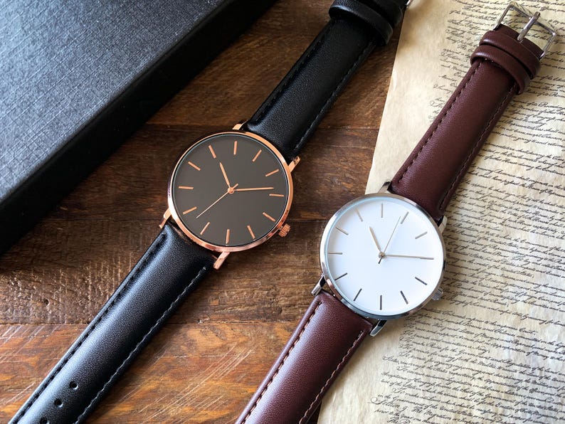 May include: Two minimalist wristwatches. One has a black face with a rose gold case and a black leather band. The other has a white face with a silver case and a brown leather band. Both have simple stick hour markers.