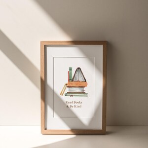 A framed print with an illustration of a stack of books and the text "Read Books & Be Kind" is displayed on a white wall, with sunlight casting shadows on the wall.
