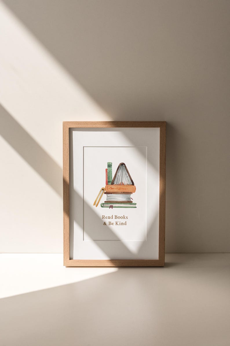 A framed print with an illustration of a stack of books and the text "Read Books & Be Kind" is displayed on a white wall, with sunlight casting shadows on the wall.