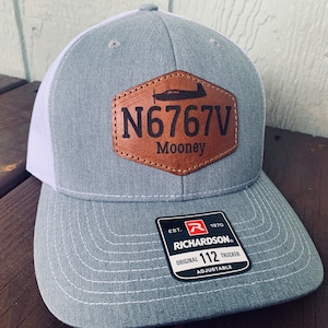 May include: Gray and white trucker hat with a brown leather patch that says "N6767V Mooney". The hat is adjustable and has a Richardson logo on the back.