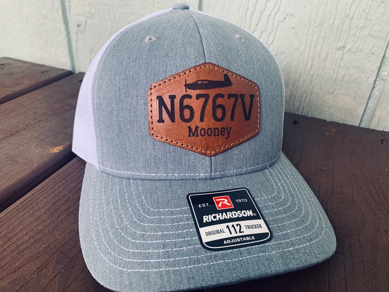May include: Gray and white trucker hat with a brown leather patch that says "N6767V Mooney". The hat is adjustable and has a Richardson logo on the back.
