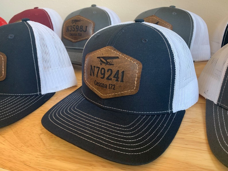 May include: A collection of five trucker hats with a white mesh back and a brown leather patch on the front. The patches feature a small airplane graphic and the text "N3598J Cessna 150" and "N79241 Cessna 172".