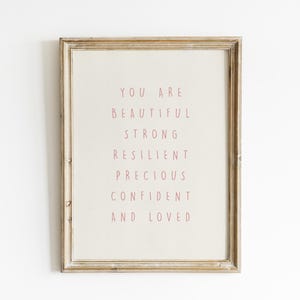 May include: A framed print with a light beige background and a distressed wooden frame. The text in the center reads "YOU ARE BEAUTIFUL STRONG RESILIENT PRECIOUS CONFIDENT AND LOVED" in a soft red color. The frame has a vintage aesthetic.