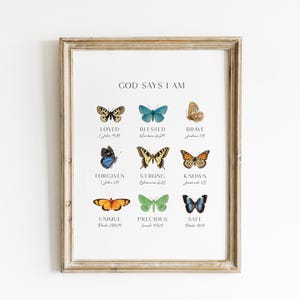 May include: A framed print with the words "GOD SAYS I AM" and colorful butterfly illustrations. Each butterfly is paired with a positive affirmation such as "LOVED" and "BLESSED". The frame is a light brown color.