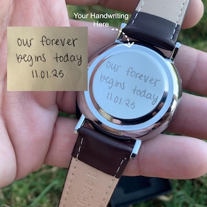May include: A silver watch with a brown leather band. The watch face has the inscription "our forever begins today 11.01.25". A small note with the same inscription is also visible.