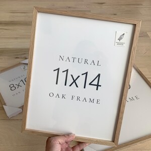 A person is holding a white picture frame with the text "NATURAL 11x14 OAK FRAME" on it.