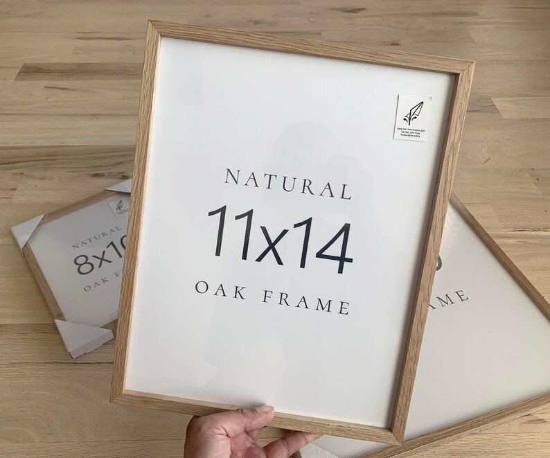 A person is holding a white picture frame with the text "NATURAL 11x14 OAK FRAME" on it.