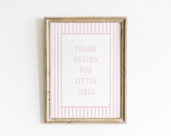 May include: A rectangular wooden frame containing a white print with vertical light pink stripes and a central border. The text in the center reads, "THANK HEAVEN FOR LITTLE GIRLS" in a simple, light pink, capitalized sans-serif font.