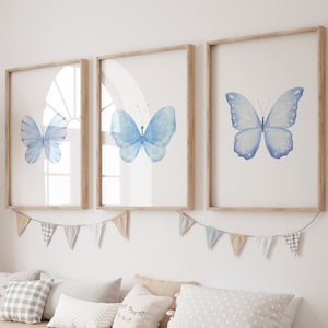 May include: Three framed prints of blue watercolor butterflies on a white background. The frames are light wood and the prints are arranged in a row.