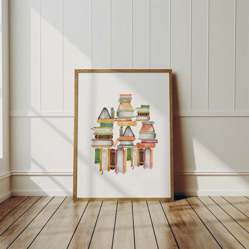 A framed watercolor painting depicting a stack of colorful books is displayed on a wooden floor against a white wall.