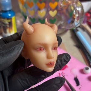 May include: A pale, smooth-skinned doll head featuring two small, curved horns protruding from the top. The doll has detailed facial features, including glossy, light red eyes and soft, natural-toned lips with a subtle sheen, presented in a neutral tone.