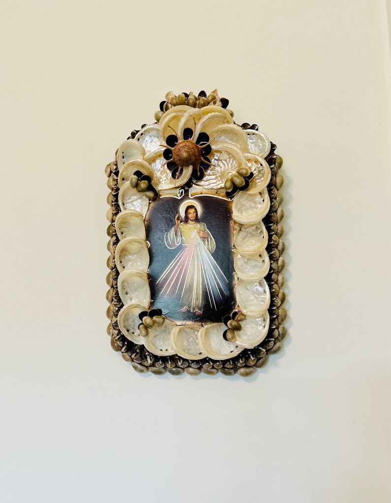 Op de afbeelding: Een ingelijst beeld van Jezus Christus met een hart en lichtstralen, omringd door een decoratief frame van schelpen.