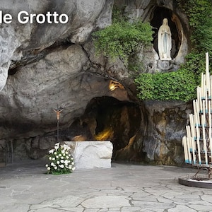 Op de afbeelding: Een wit beeld van de Maagd Maria staat in een grot, met een grote kandelaar op de voorgrond. De grot heet Massabielle Grotto.
