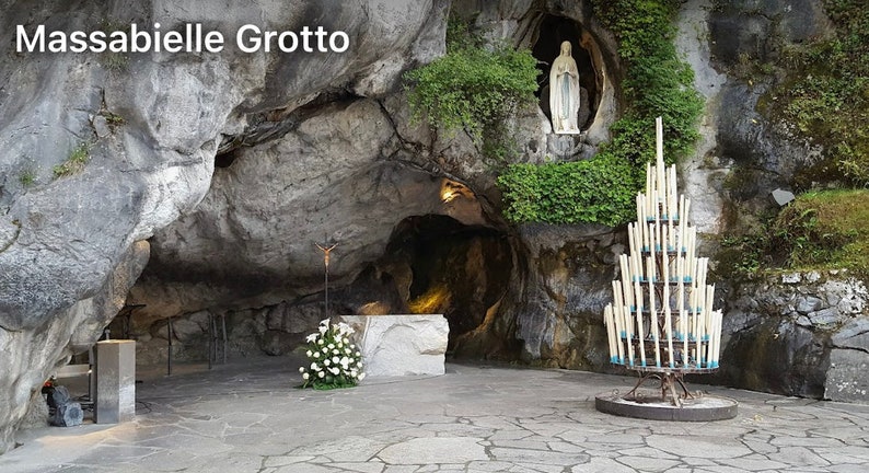 Op de afbeelding: Een wit beeld van de Maagd Maria staat in een grot, met een grote kandelaar op de voorgrond. De grot heet Massabielle Grotto.