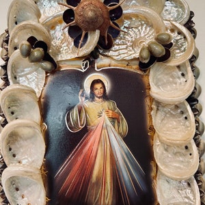 Op de afbeelding: Een ingelijst beeld van Jezus Christus met een rood, geel en blauw stralend licht achter hem. De lijst is gemaakt van schelpen en andere natuurlijke materialen.