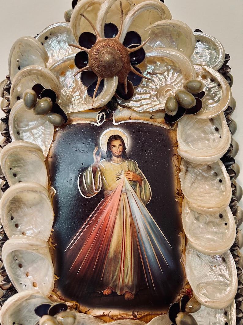 Op de afbeelding: Een ingelijst beeld van Jezus Christus met een rood, geel en blauw stralend licht achter hem. De lijst is gemaakt van schelpen en andere natuurlijke materialen.