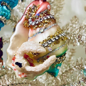 May include: A decorative Christmas ornament shaped like a seashell. The ornament is primarily pink, white, and teal, with gold and silver embellishments. It features intricate details and is adorned with rhinestones and beads.