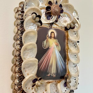 Op de afbeelding: Een ingelijst beeld van Jezus Christus met een hart en lichtstralen erachter. De lijst is gemaakt van schelpen en andere natuurlijke materialen.