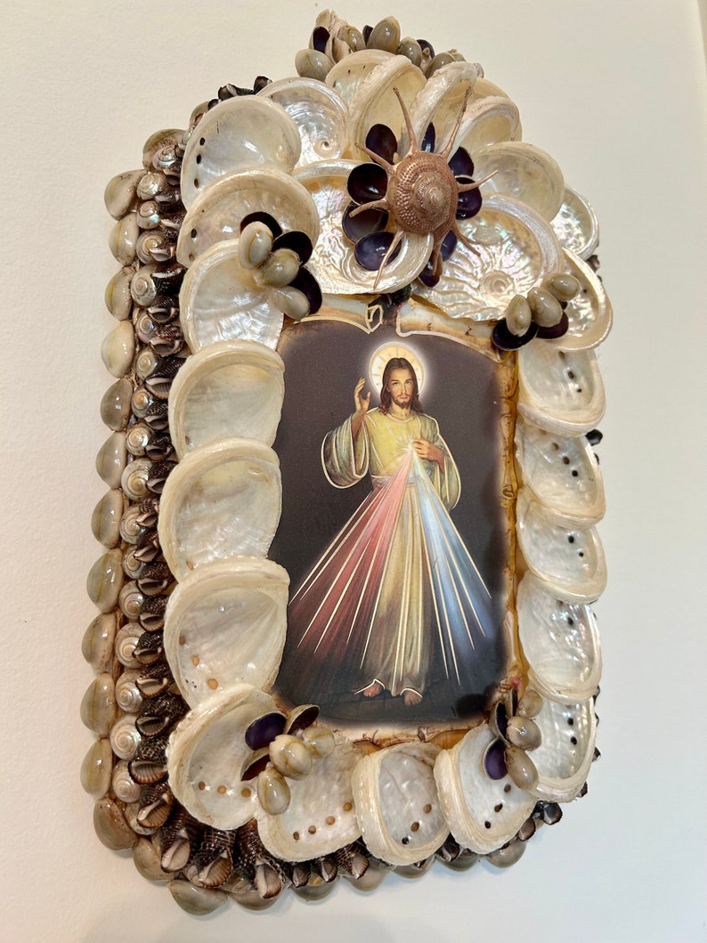 Op de afbeelding: Een ingelijst beeld van Jezus Christus met een hart en lichtstralen erachter. De lijst is gemaakt van schelpen en andere natuurlijke materialen.