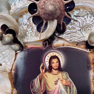 Op de afbeelding: Een ingelijst beeld van Jezus Christus met een halo en een rood, geel en blauw hart. De lijst is versierd met schelpen en een zeester.