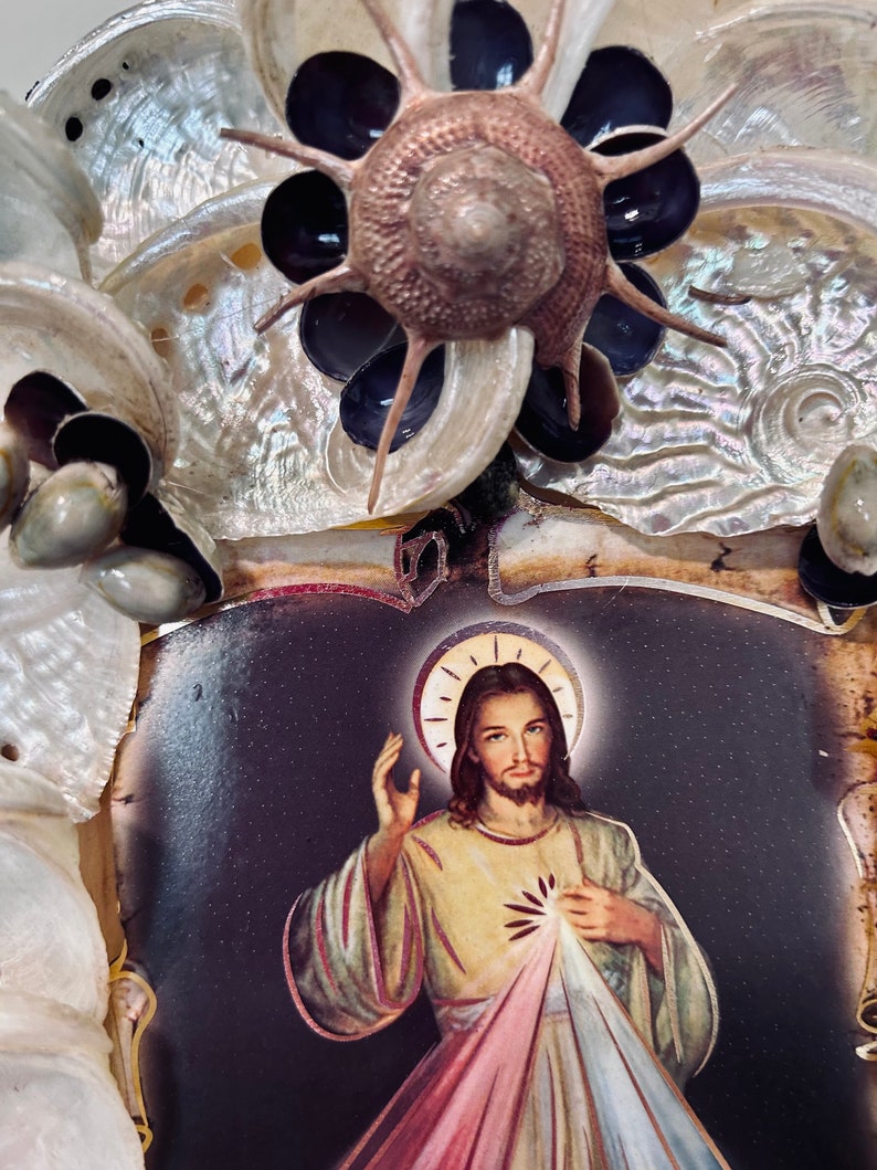 Op de afbeelding: Een ingelijst beeld van Jezus Christus met een halo en een rood, geel en blauw hart. De lijst is versierd met schelpen en een zeester.