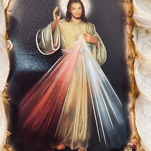 Op de afbeelding: Een ingelijst beeld van Jezus Christus met een halo en lichtstralen die uit zijn borst komen. Het beeld is geplaatst tegen een donkere achtergrond met een sterreneffect. Het beeld is ingelijst met een decoratieve rand van schelpen.