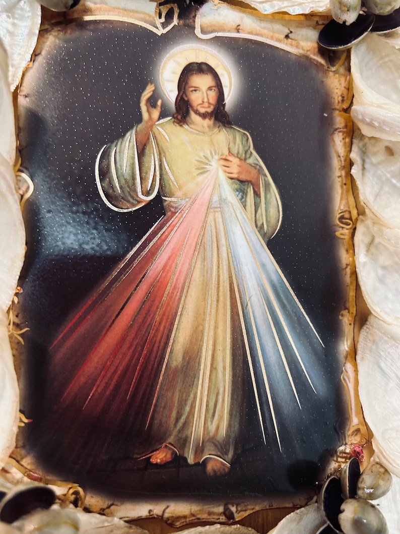 Op de afbeelding: Een ingelijst beeld van Jezus Christus met een halo en lichtstralen die uit zijn borst komen. Het beeld is geplaatst tegen een donkere achtergrond met een sterreneffect. Het beeld is ingelijst met een decoratieve rand van schelpen.