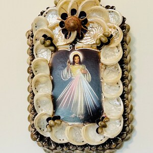 Op de afbeelding: Een ingelijst beeld van Jezus Christus met een hart en lichtstralen, omringd door een decoratief frame van schelpen.