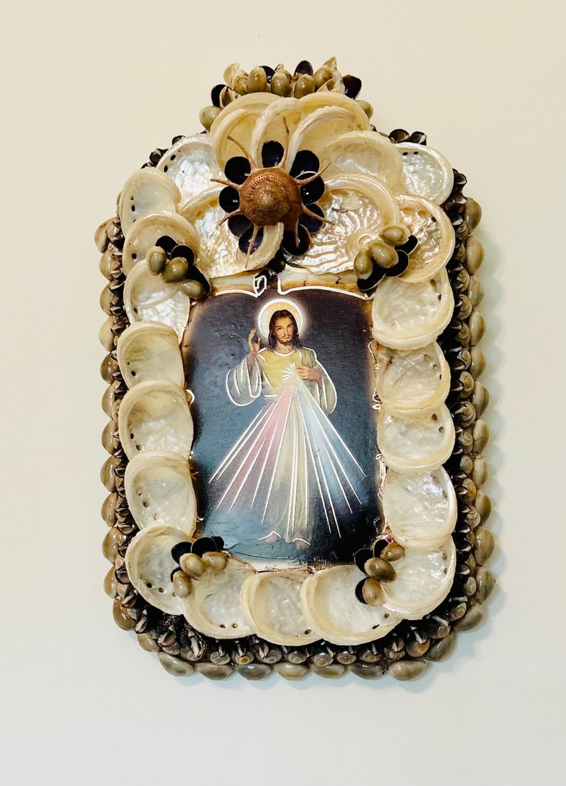 Op de afbeelding: Een ingelijst beeld van Jezus Christus met een hart en lichtstralen, omringd door een decoratief frame van schelpen.