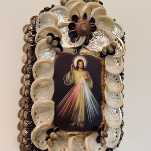 Op de afbeelding: Een ingelijst beeld van Jezus Christus met een hart van vuur en lichtstralen, omringd door een decoratief frame van schelpen.