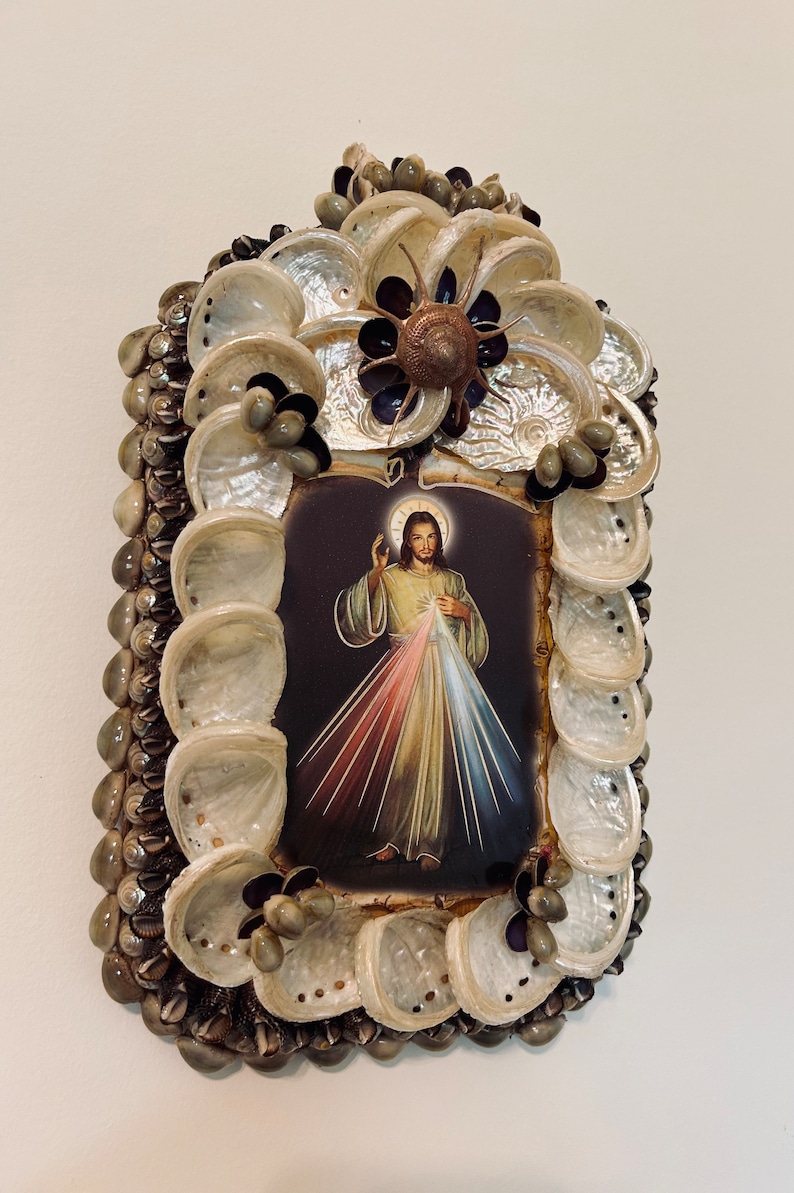 Op de afbeelding: Een ingelijst beeld van Jezus Christus met een hart van vuur en lichtstralen, omringd door een decoratief frame van schelpen.