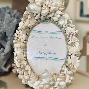 May include: Oval picture frame decorated with various seashells in shades of white and cream. The frame features the text "Shell & Sea Creations by Sherry Josey" in blue script on a white background.