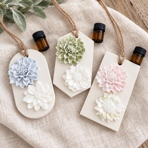 May include: Three hanging air freshener plaques in oval, hexagonal, and rectangular shapes. Each plaque is off-white and features two raised floral embellishments in pastel blue, white, sage green, or pink. Each includes a small amber glass bottle.