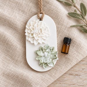 May include: An oval white hanging diffuser plaque featuring a solid white sculpted dahlia flower and a sage green marbled sculpted flower, suspended by a jute twine cord, accompanied by a small amber glass essential oil bottle.
