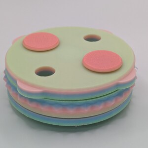 May include: A stack of colorful coasters. The top coaster is light green with three small holes and three pink circles. The coasters below are pink, blue, and light green. The coasters are round with a wavy edge.