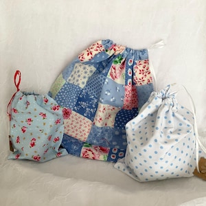 May include: Three drawstring bags in different sizes and patterns. The largest bag has a patchwork design in blue and pink hues. The smaller bags feature floral and polka dot patterns. All bags have white drawstrings.