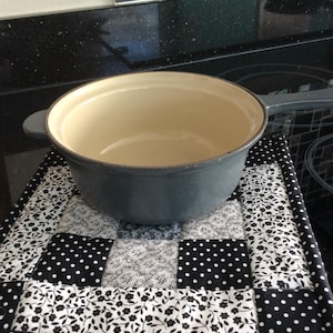 May include: A dark grey cast iron pot with a cream interior rests on a black and white quilted hot pad. The hot pad has a patchwork design with polka dots and floral patterns.