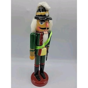 May include: A wooden nutcracker soldier in a green and red uniform with a black hat and white hair. The nutcracker has a yellow sash and is standing on a red base. The figure is approximately 12 inches tall.