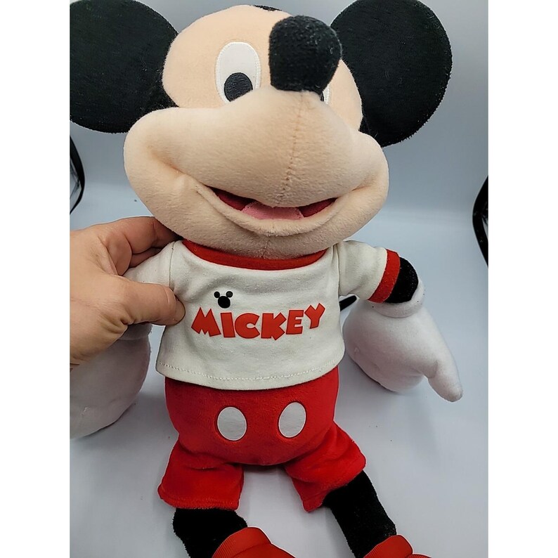 May include: A plush Mickey Mouse doll with a cream-colored face, black ears, and a red and white outfit. The white shirt has the word "Mickey" in red. The doll is approximately 46 cm tall.