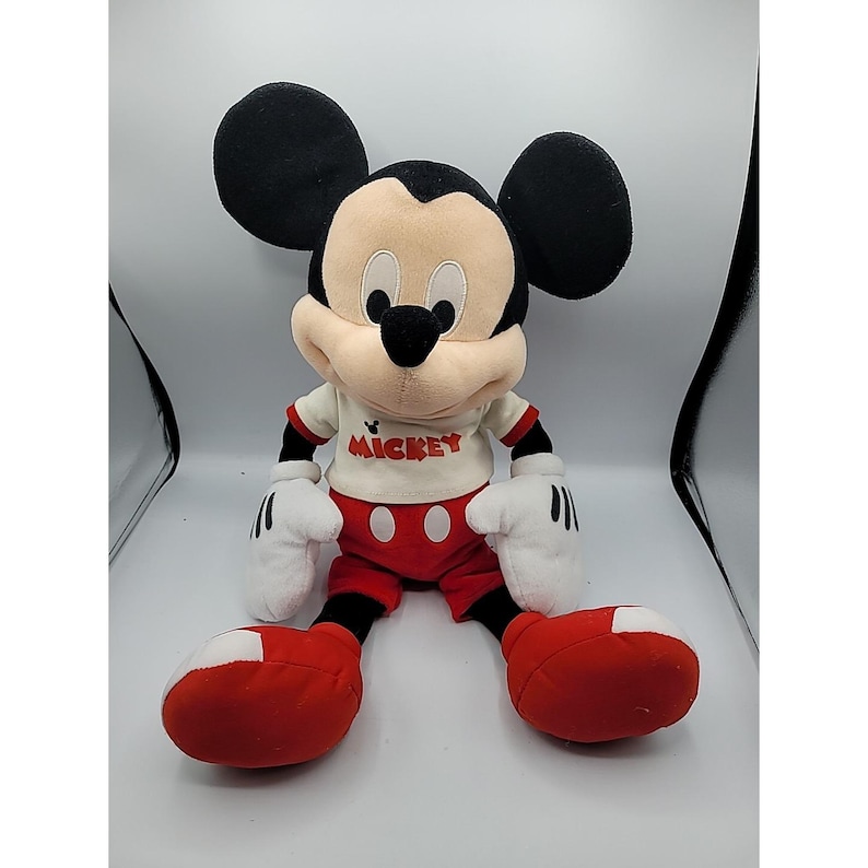 May include: A plush Mickey Mouse doll, with a cream-coloured face, black ears, and a black nose. Mickey wears a white shirt with red trim and the word "Mickey" printed on it, red shorts, and red shoes. The doll has white gloves.