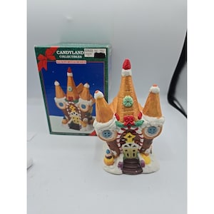 May include: A Candyland Collectibles porcelain lighted house with a gingerbread design. The house features orange, white, and brown colours, with red and green accents. The box behind the house has the Candyland Collectibles logo and a picture of the house.