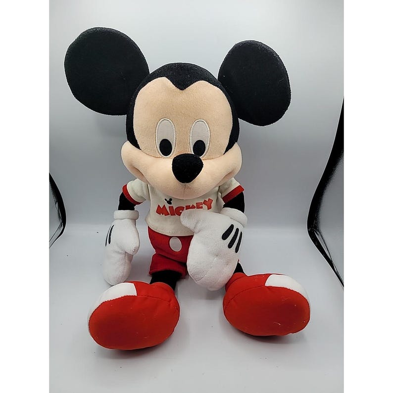 May include: A soft toy of Mickey Mouse, featuring large black ears, a cream-coloured face, and a black nose. Mickey is dressed in a white shirt with "Mickey" printed on it, red shorts, and large red shoes. His white gloves have black lines.