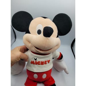 May include: A Mickey Mouse soft toy with black ears, a beige face, and a white top with the word "Mickey" in red. The toy has red shorts and black eyes. The toy is a classic Disney character.