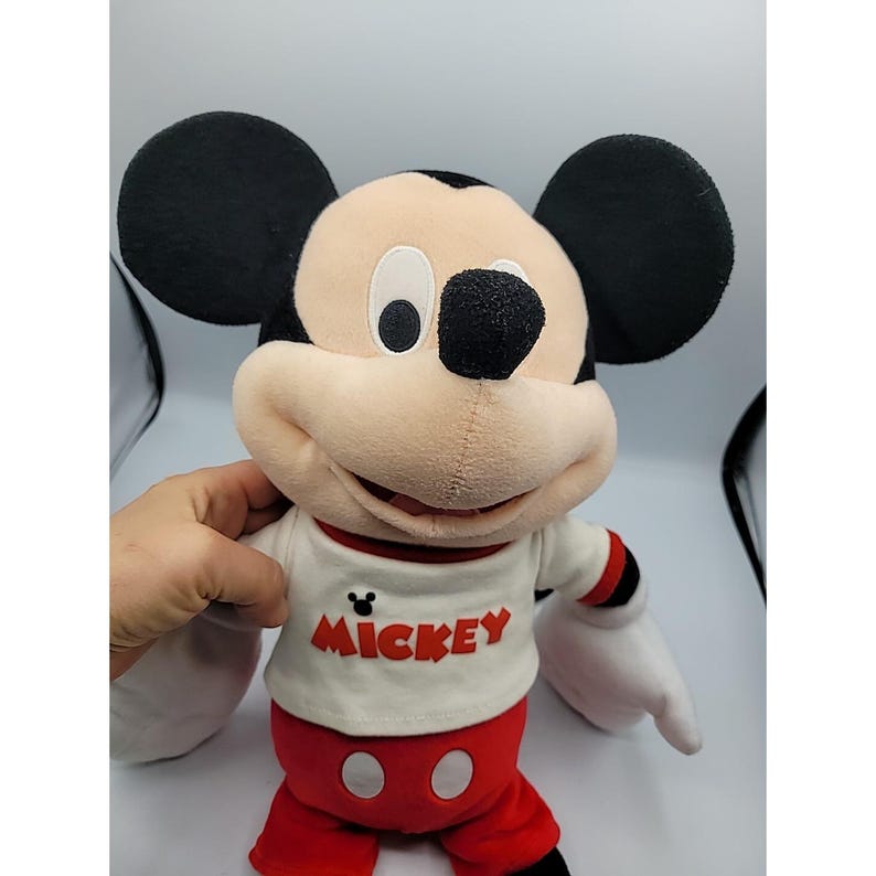 May include: A Mickey Mouse soft toy with black ears, a beige face, and a white top with the word "Mickey" in red. The toy has red shorts and black eyes. The toy is a classic Disney character.