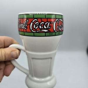 Könnte beinhalten: Weiße Keramiktasse in Form eines Coca-Cola-Glases mit einem rot-grün-weißen Buntglasdesign mit dem Text "Drink Coca-Cola".