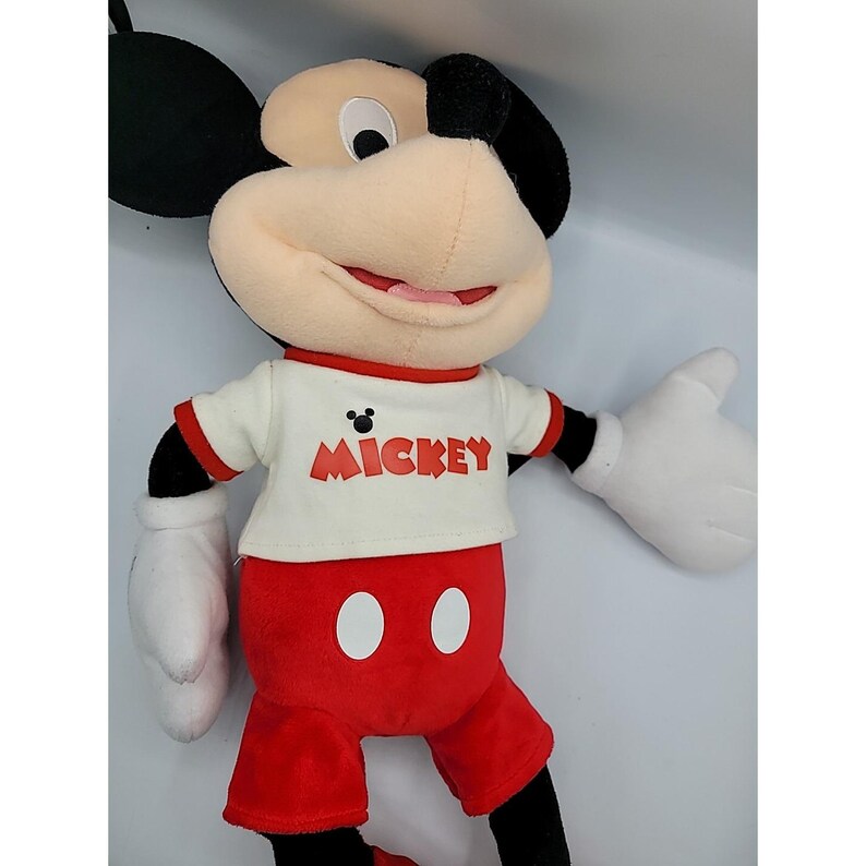 May include: A plush Mickey Mouse doll. Mickey is wearing a white t-shirt with red trim and the word "Mickey" printed on the front. He has red shorts, white gloves, and black ears. The doll is approximately 46 cm tall.