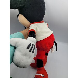 May include: A plush Mickey Mouse doll, with a black head and ears, a white shirt with red trim, red shorts, and large white gloves with black details. The doll has a black tail and red shoes.