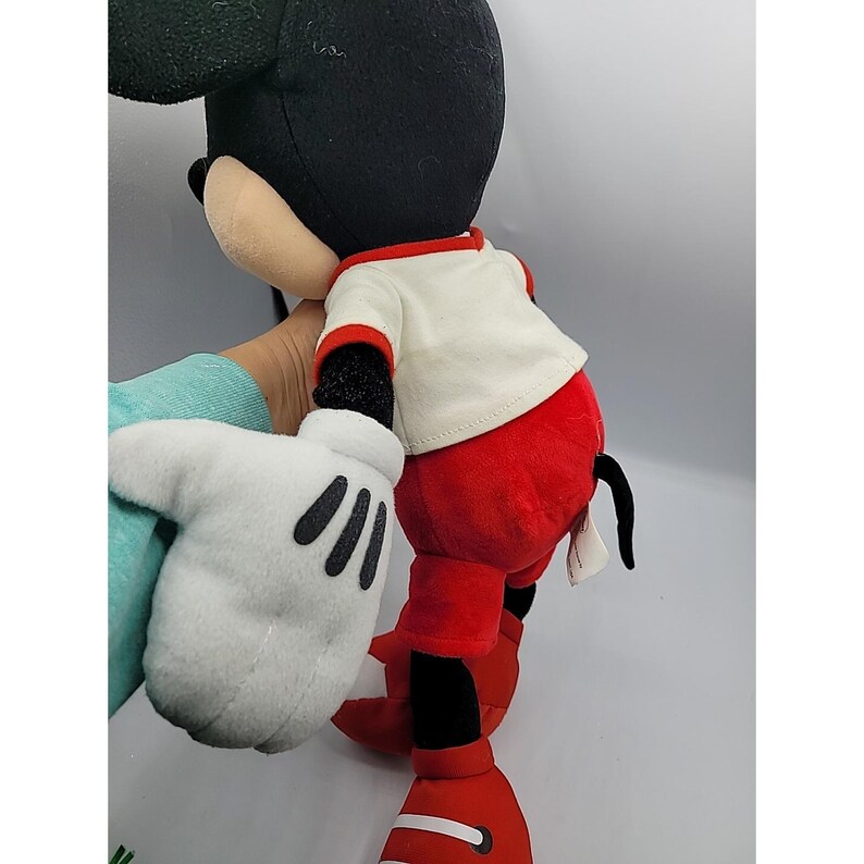 May include: A plush Mickey Mouse doll, with a black head and ears, a white shirt with red trim, red shorts, and large white gloves with black details. The doll has a black tail and red shoes.